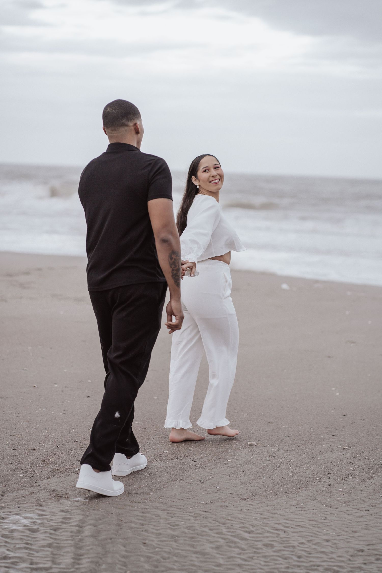 A couple in black and white outfits walking together on a misty beach shoreline.