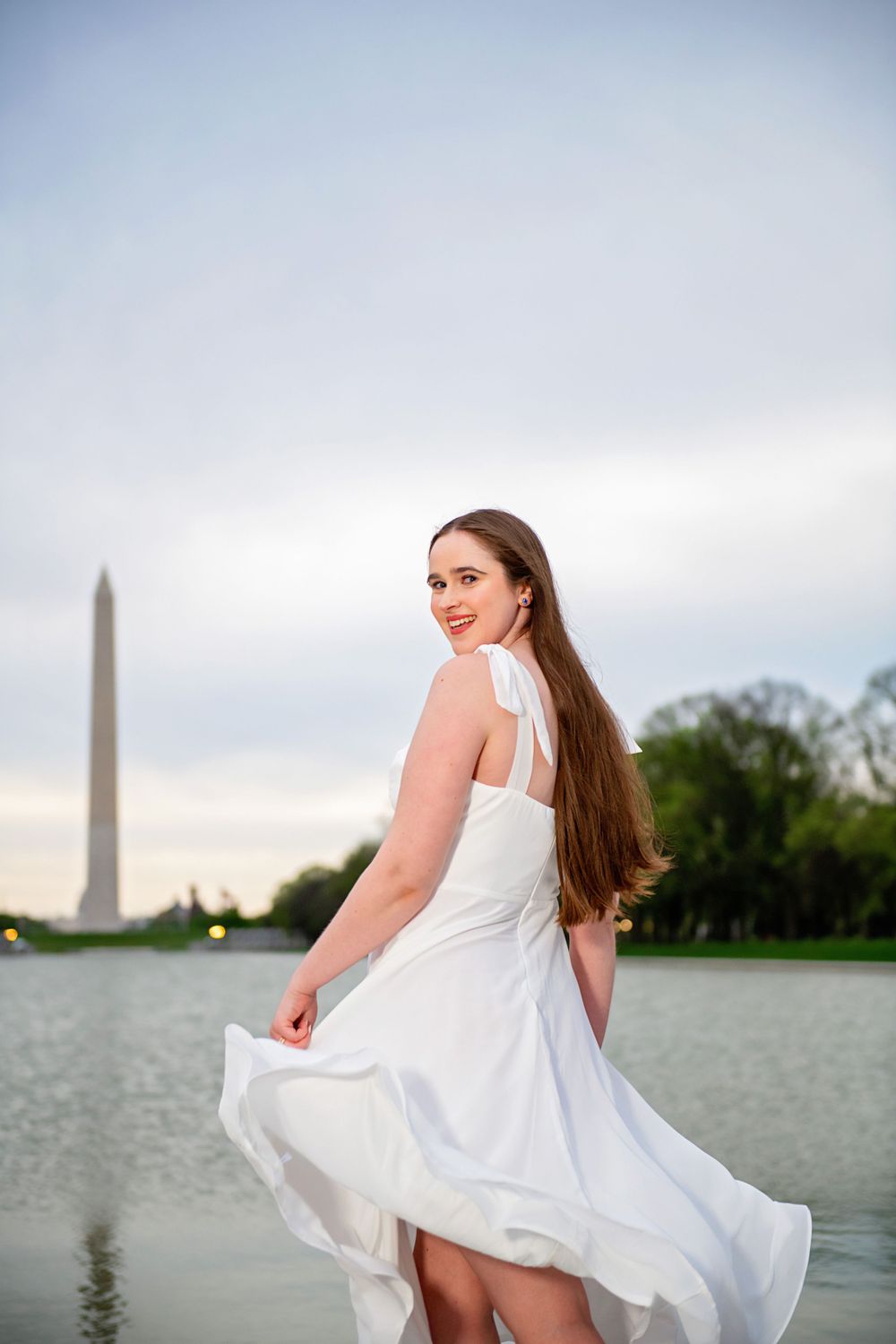 Woman in flowing white dress poses by Washington Monument reflection pool at sunset.