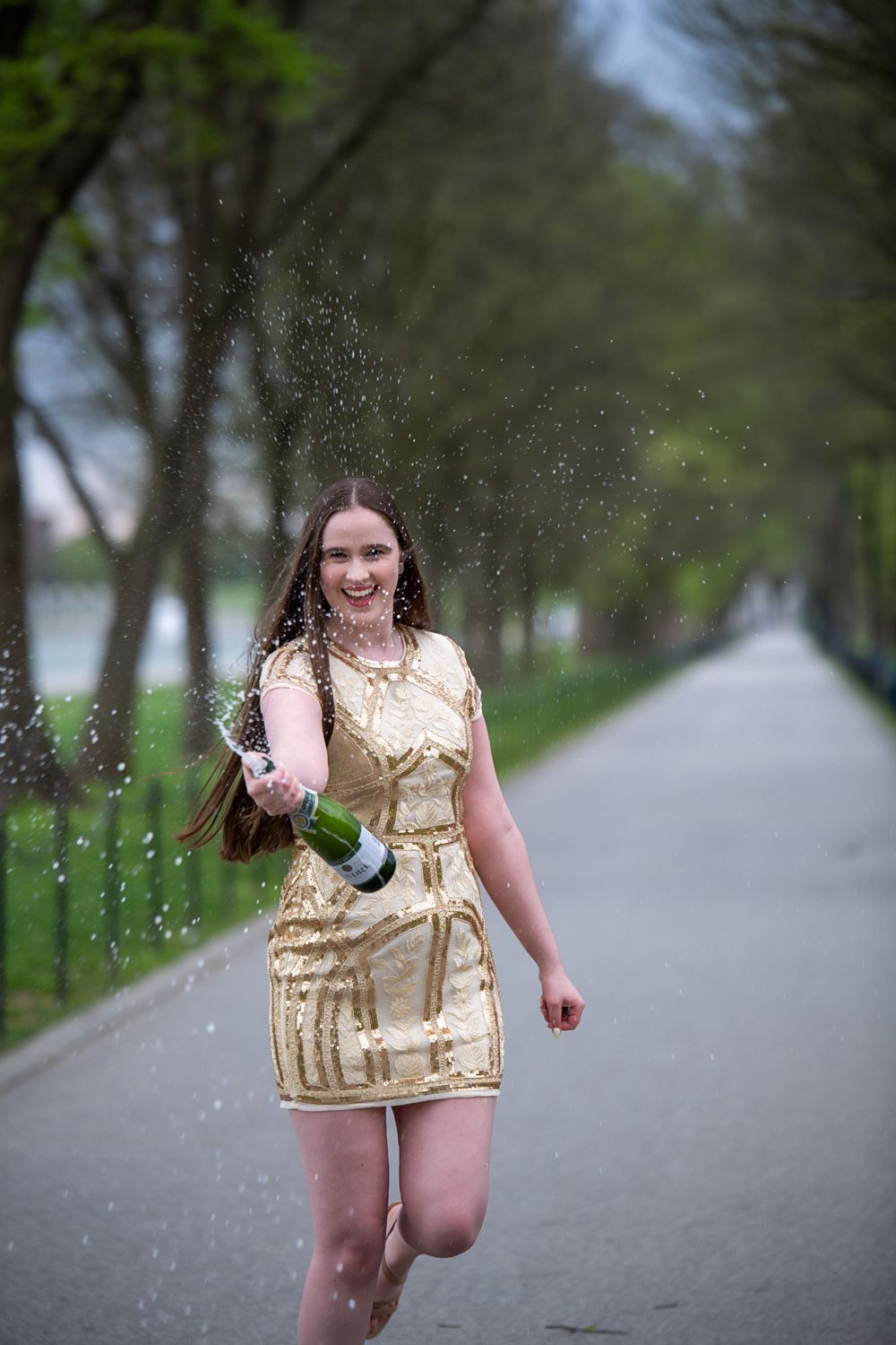 Celebratory moment with champagne spray on tree-lined path in gold sequin dress.