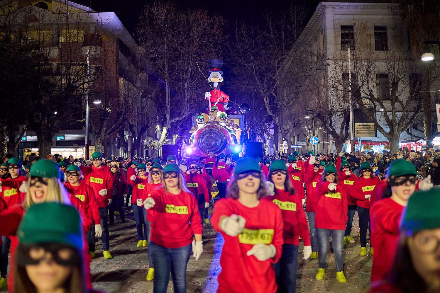 Holiday parade featuring crowd in matching red shirts and green hats gathering under illuminated display.