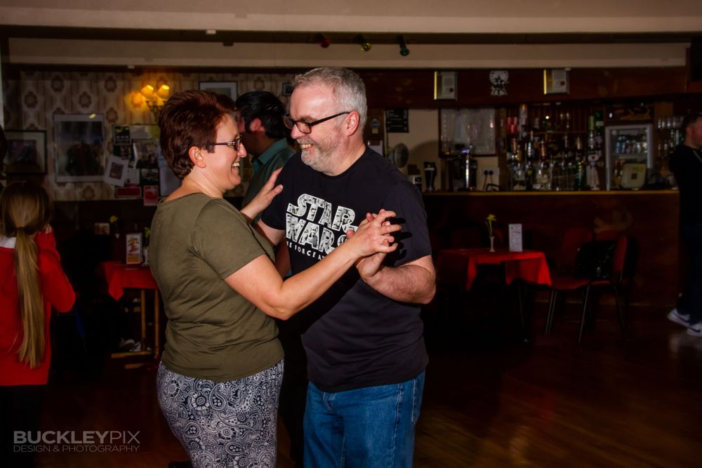 Couples dance together in a dimly lit ballroom with hardwood floors and warm lighting.