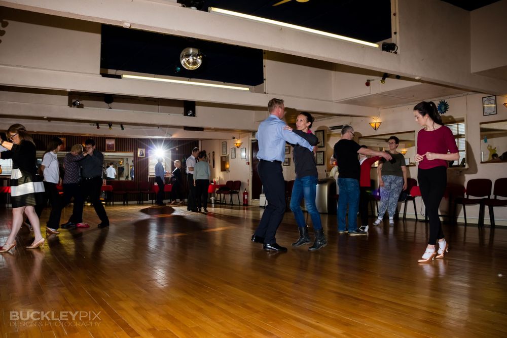 Wide angle view of dancers practicing in a large studio with wooden flooring.