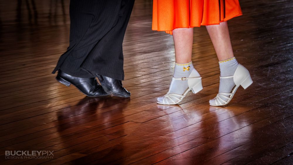 Close-up of dancers' feet in various shoes moving across a polished wooden dance floor.