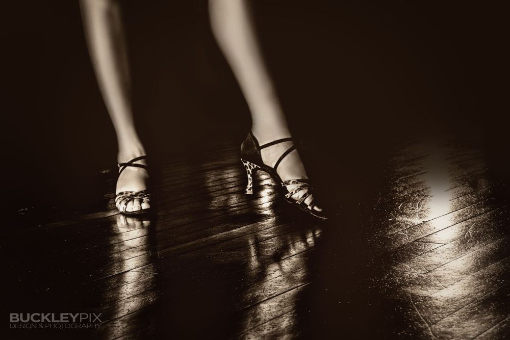 Monochrome image of a female dancer's legs from the knees down, wearing dance shoes on a shiny dance floor.