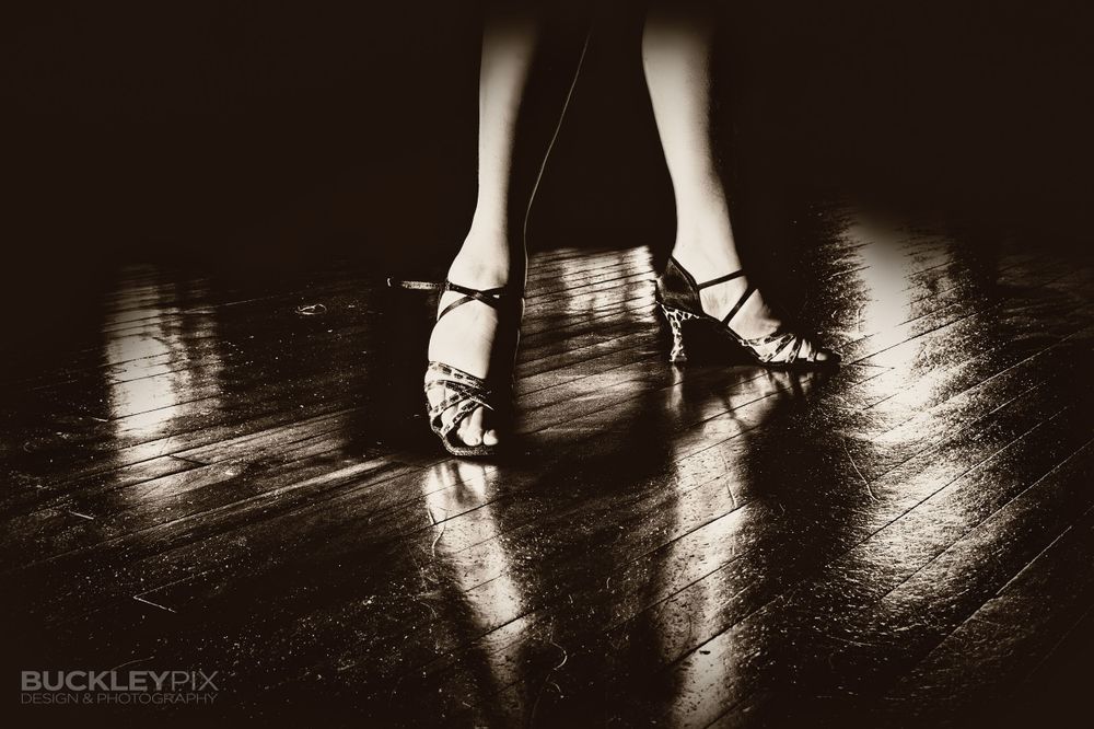 Dramatic low-angle silhouette of dancers' feet on a reflective wooden floor.