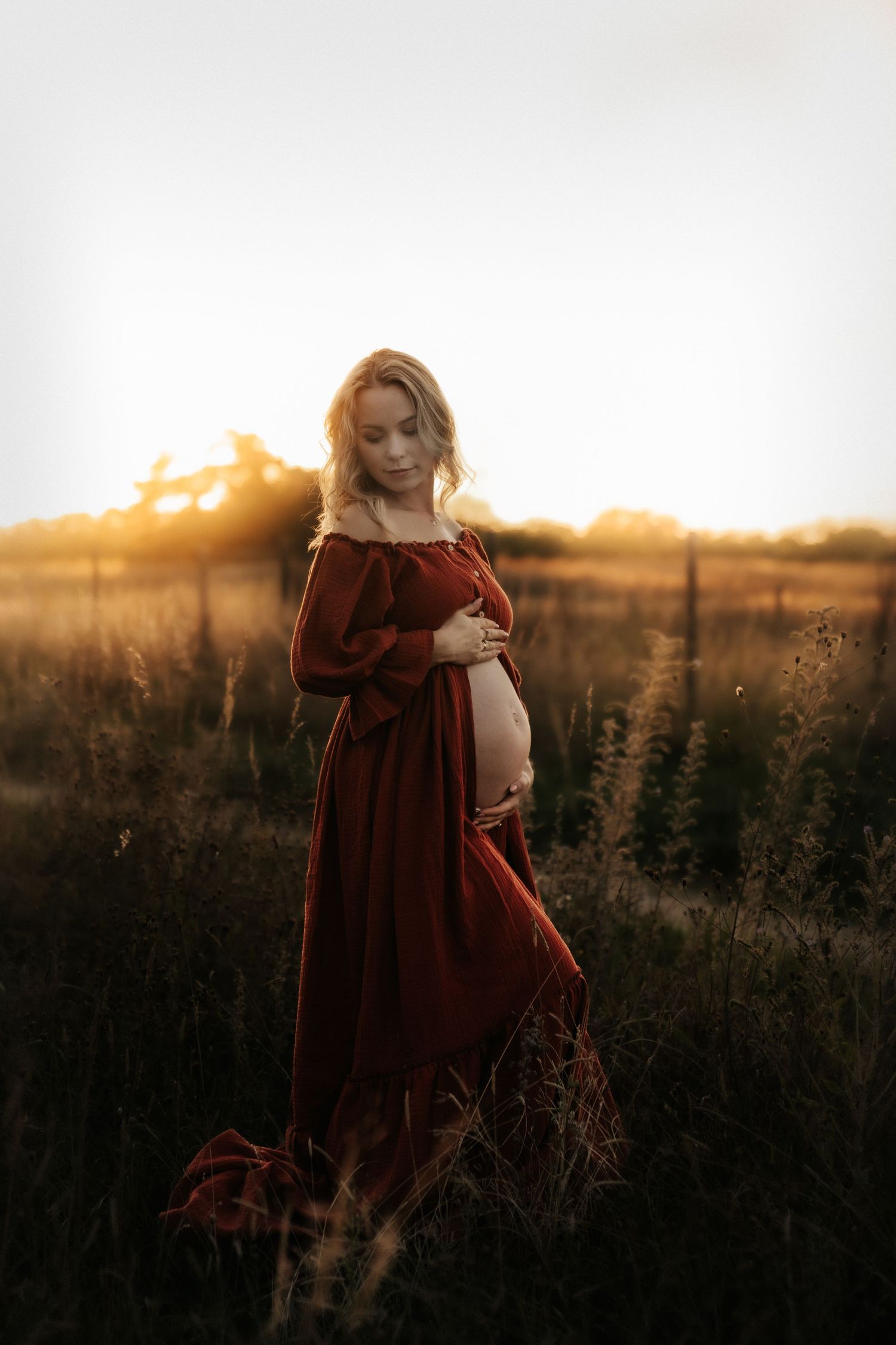 A person in a flowing burgundy dress cradles their pregnant belly in a sunlit field during golden hour.