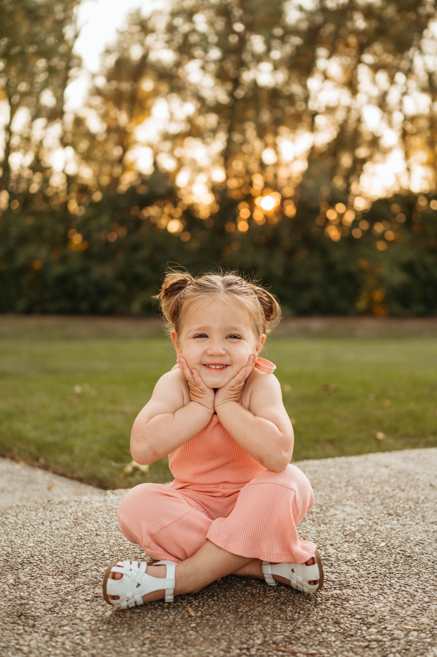 Happy toddler girl in pink romper sitting cross-legged on path with golden sunset bokeh background.