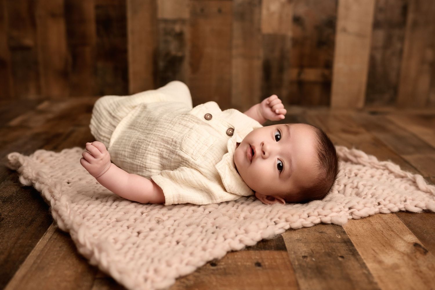 Alert newborn baby in cream knit romper lies on pink blanket over rustic wood floor backdrop.