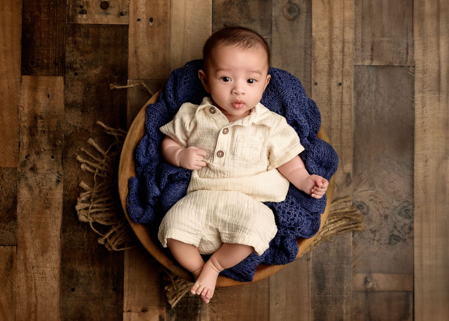 Smiling baby boy in cream romper sitting in wooden bowl with blue knit blanket.
