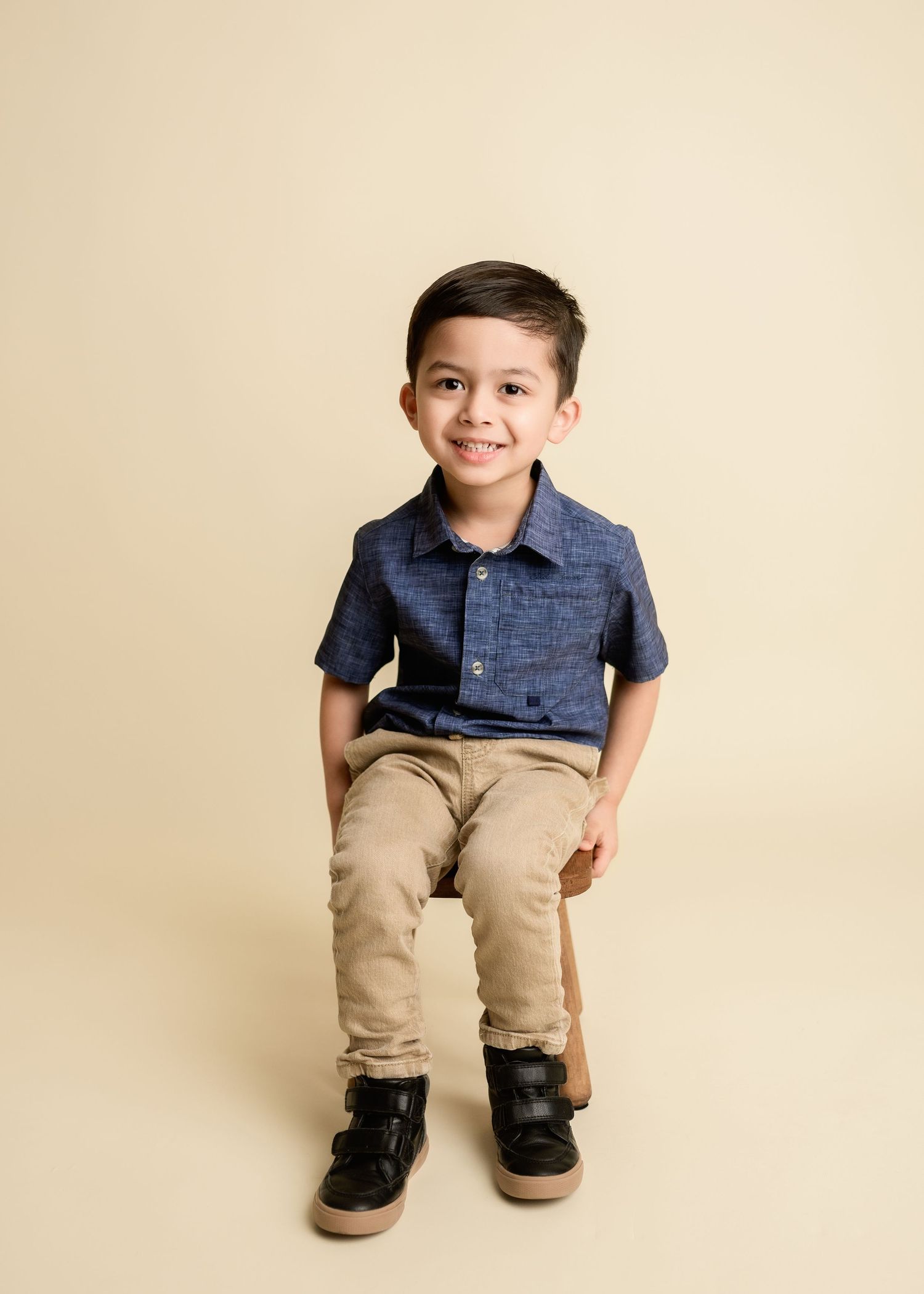 Smiling young boy in navy blue shirt and khaki pants sitting for a studio portrait photo.