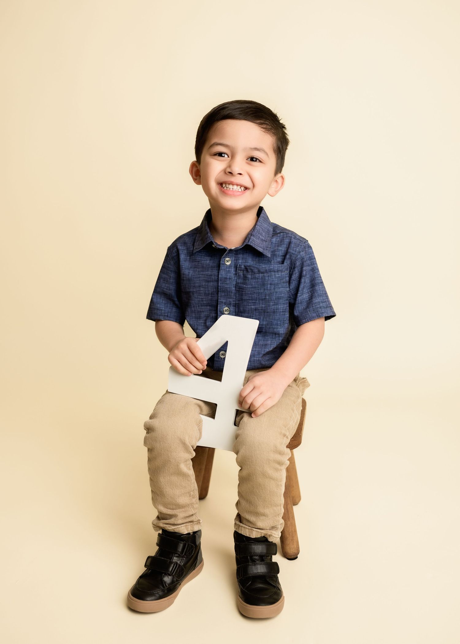 Happy young boy holding number four sign sitting in studio portrait wearing navy shirt and khakis.