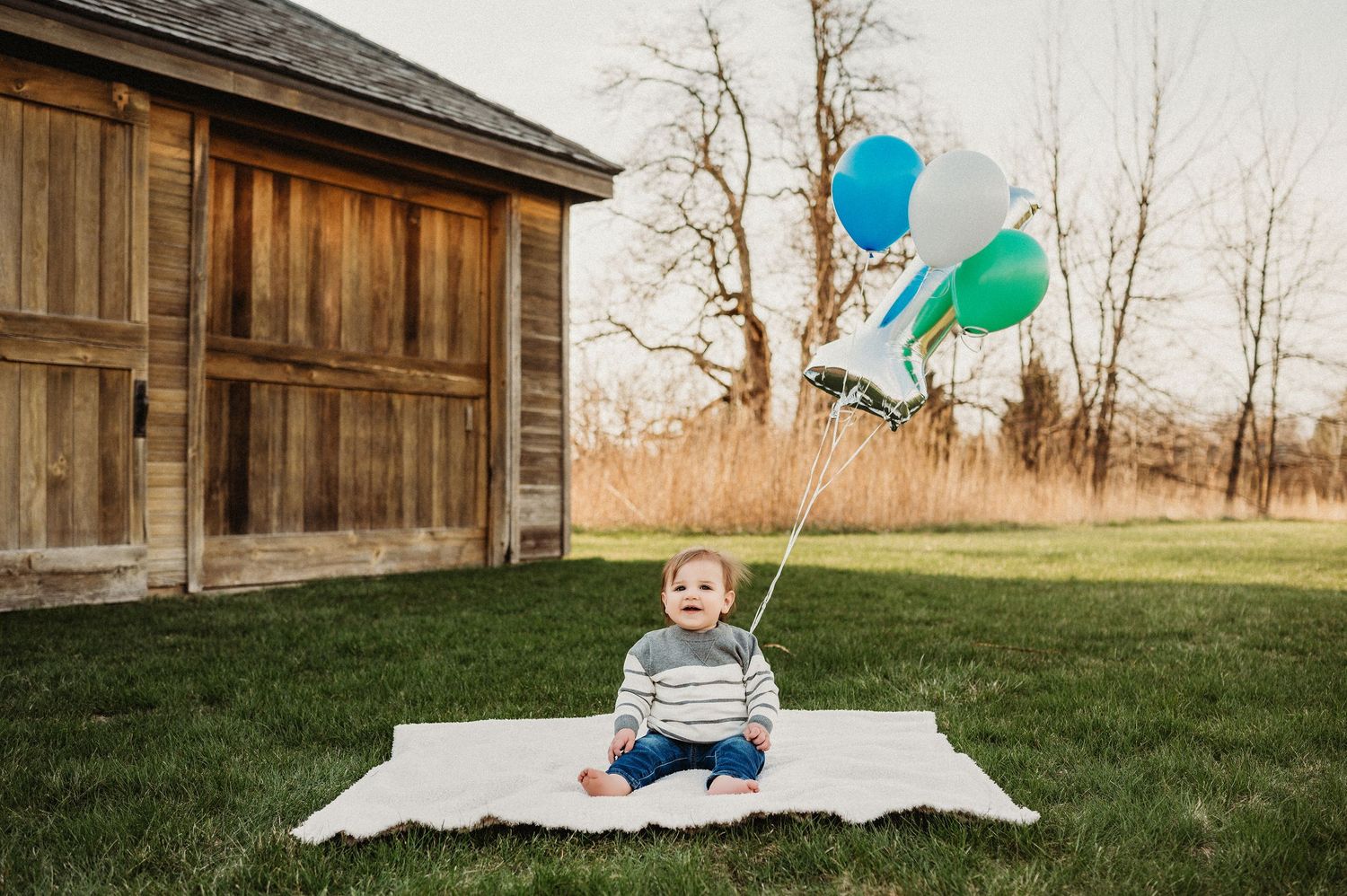 One year old baby sitting on white blanket holding colorful balloons outdoors near rustic barn.