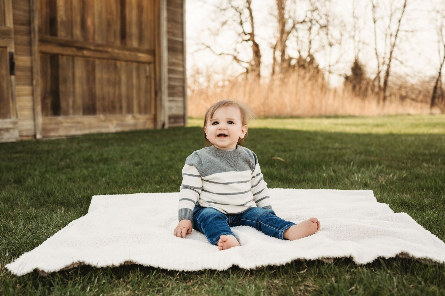 Happy toddler boy in striped sweater sitting on white blanket on green grass outdoors.