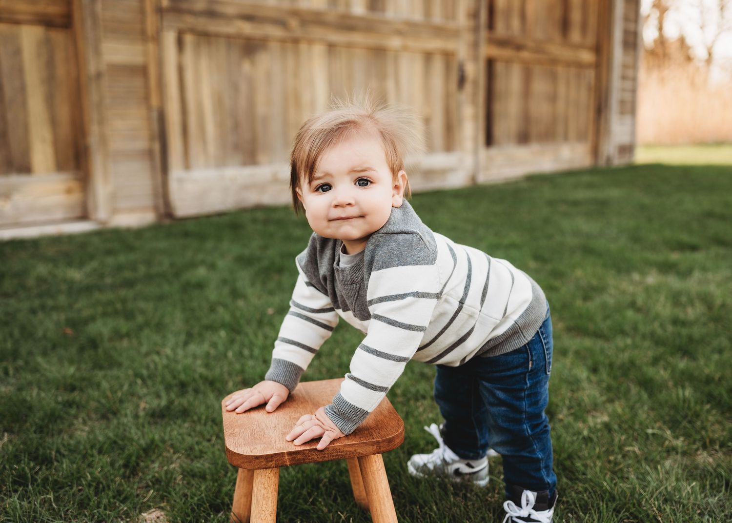 Toddler boy in striped sweater leaning on wooden stool outdoors near rustic barn.