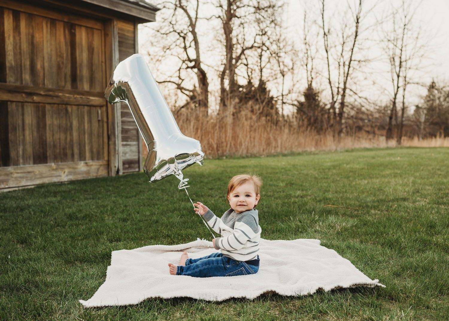 Baby sitting on blanket outdoors holding a number one balloon during first birthday photo session.