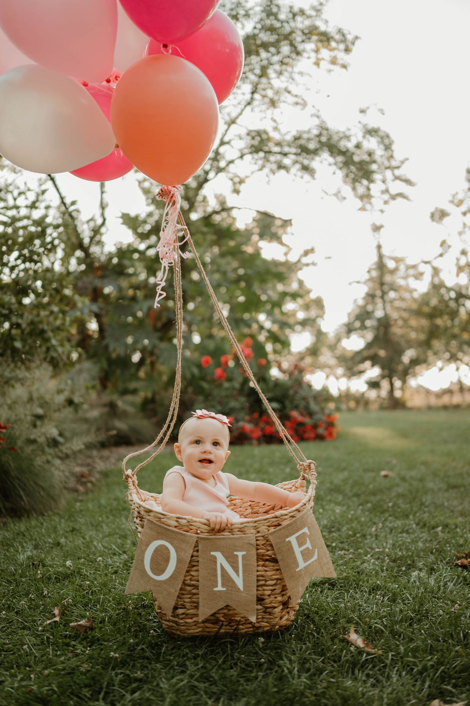 Baby girl sitting in wicker basket with pink balloons spelling ONE.