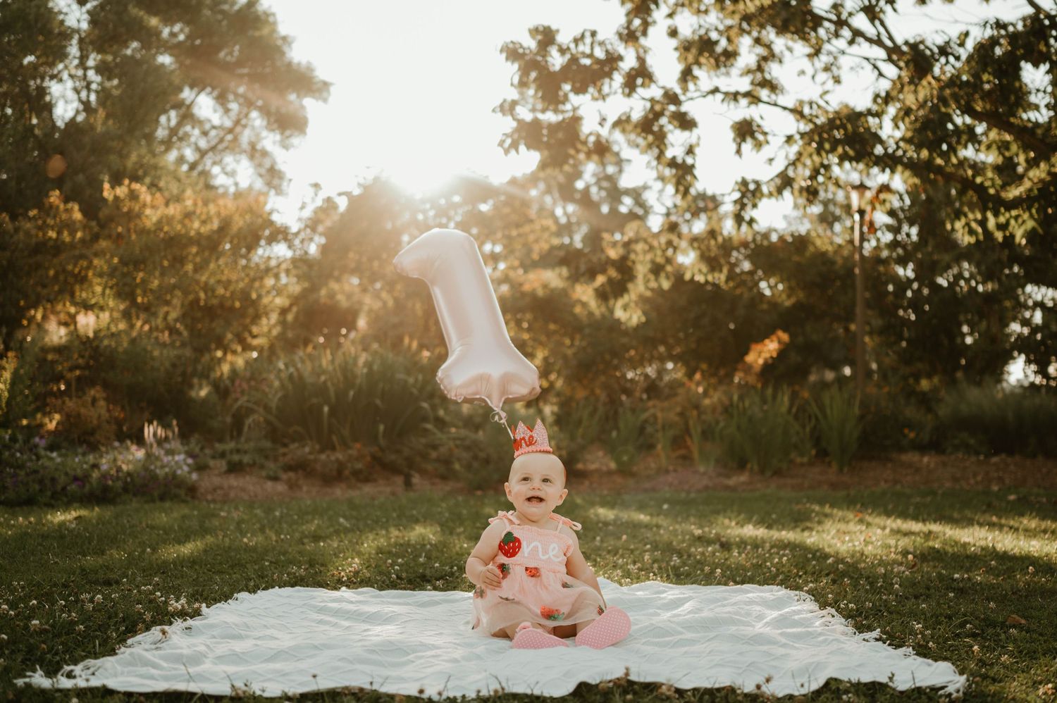 Baby girl in strawberry dress with number one balloon outdoors.
