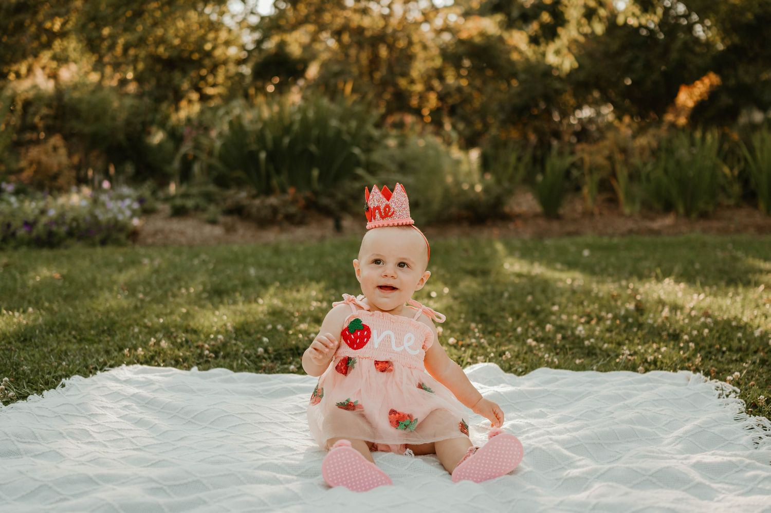 Baby girl in strawberry dress and crown sitting on white blanket.