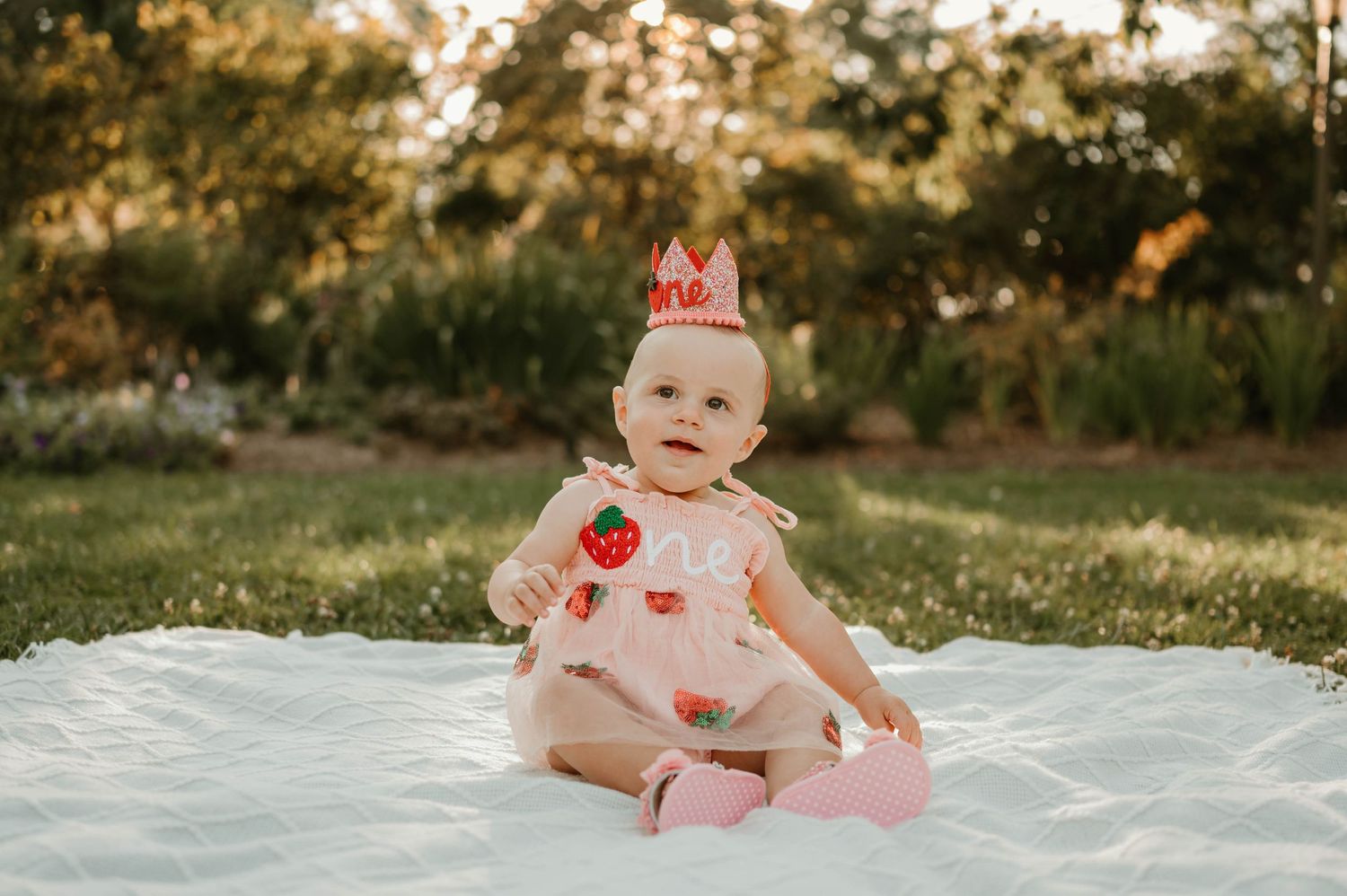 Smiling baby girl in strawberry dress and crown on white blanket.