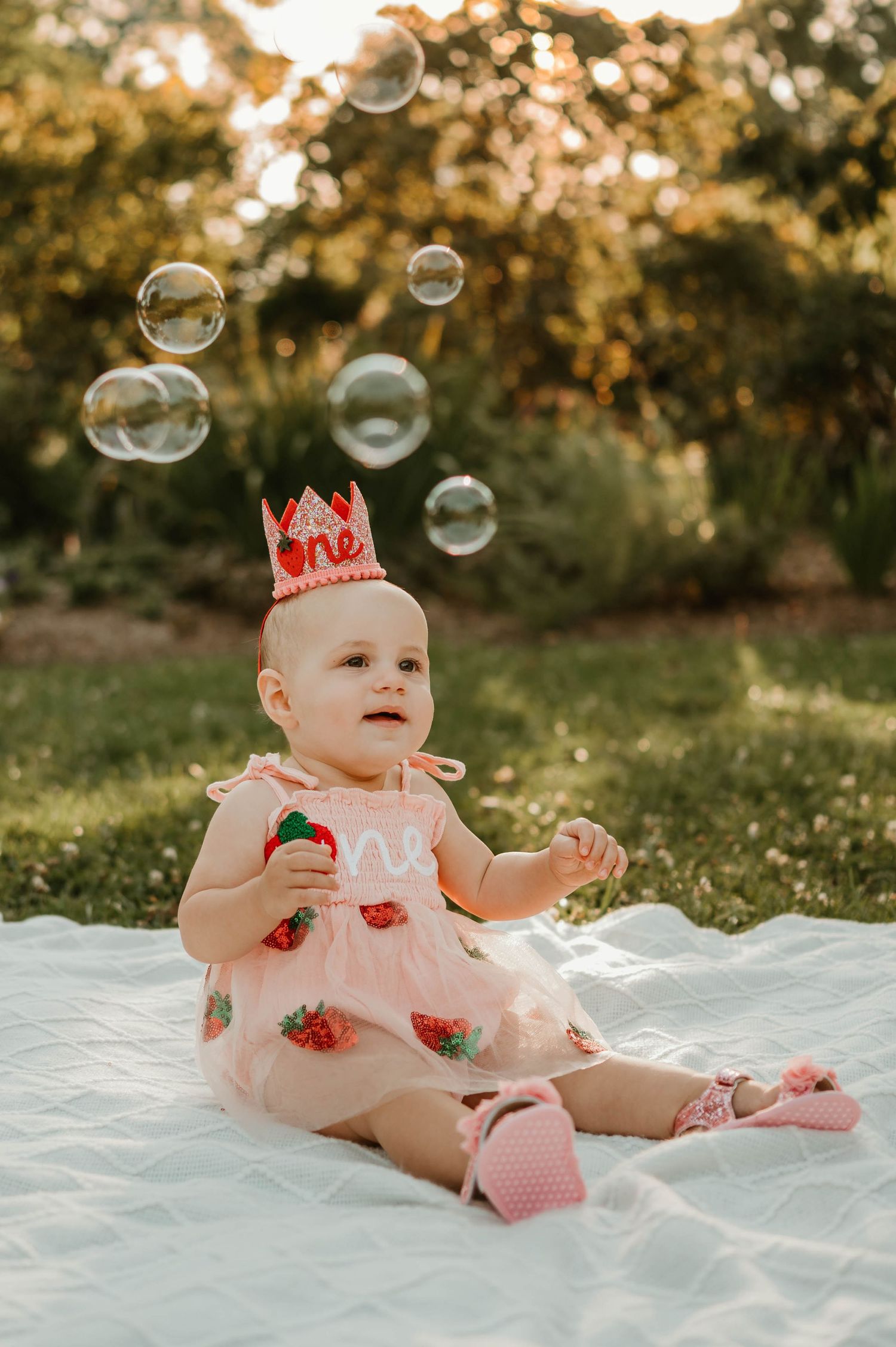 Baby girl in strawberry outfit watching bubbles float in sunny garden.