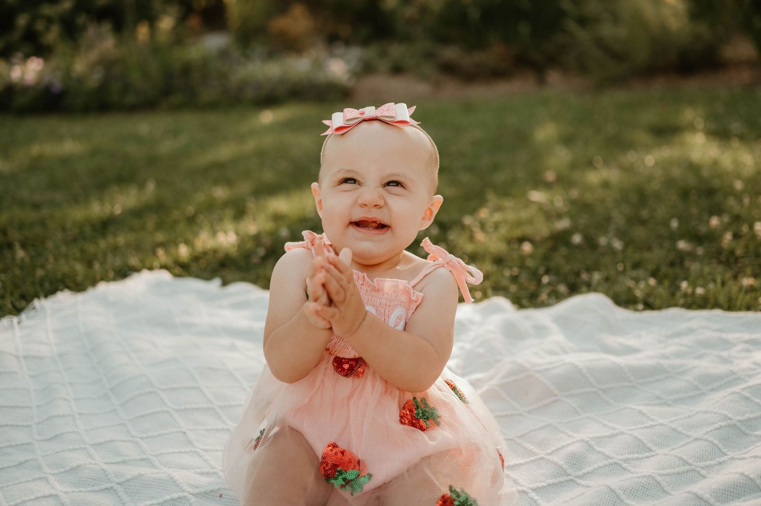 Smiling baby girl in pink floral romper clapping hands while sitting on white blanket in green grass.