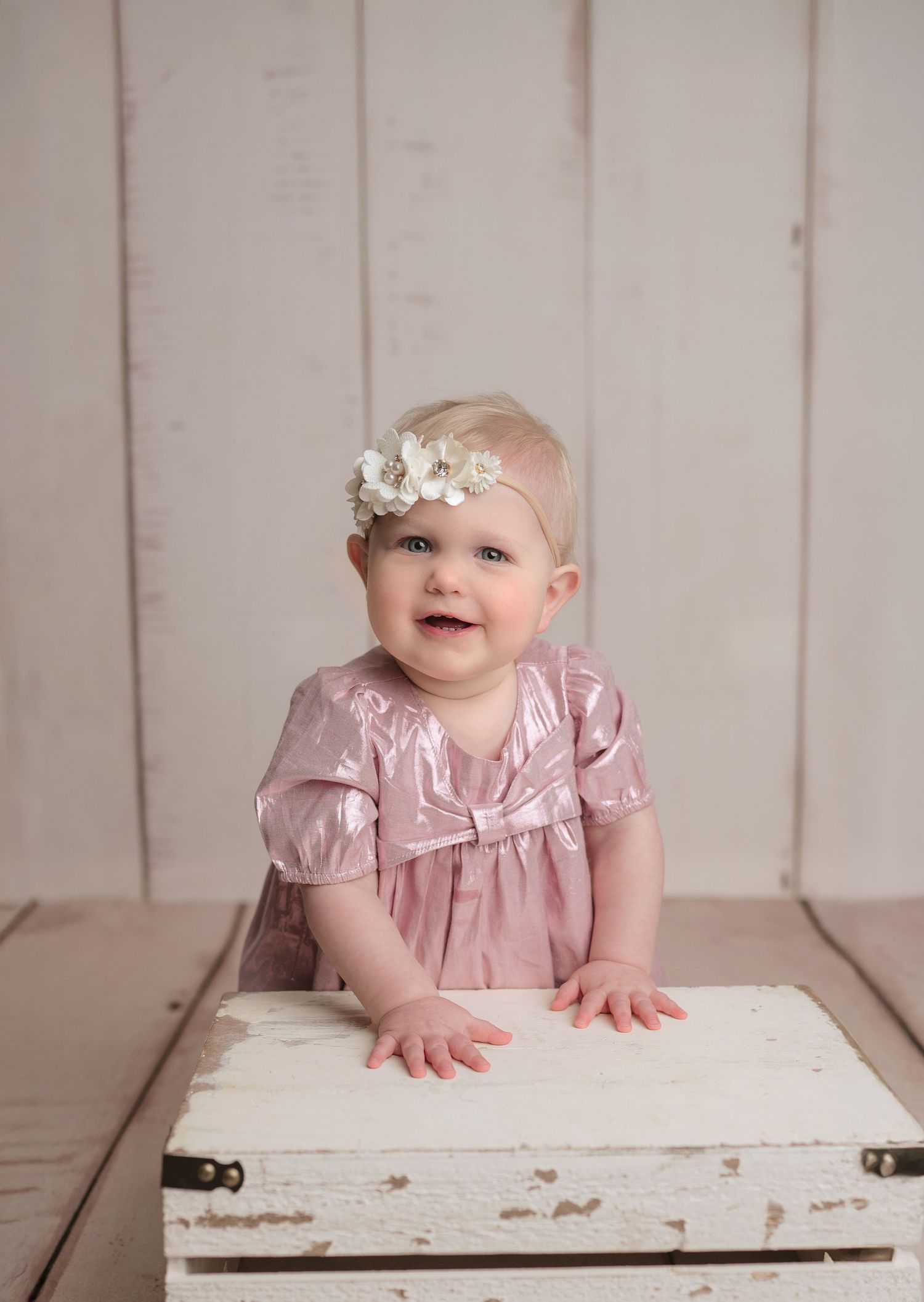 Adorable baby girl in pink velvet dress with white floral headband smiling while leaning on white box.