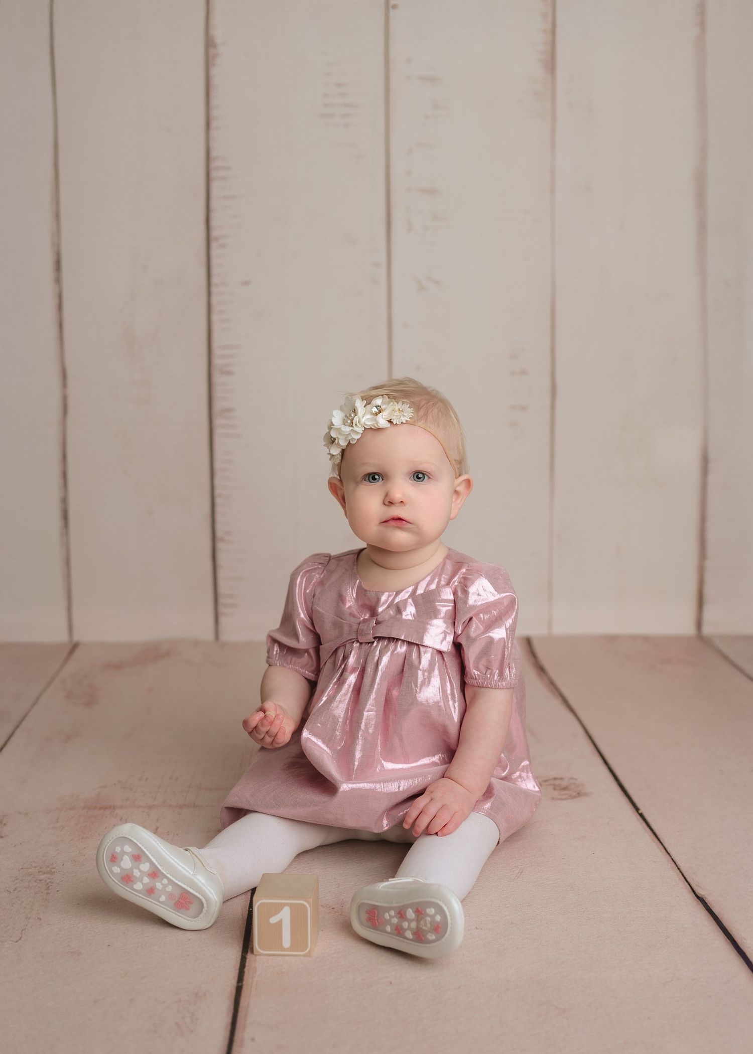 Baby girl in pink metallic dress and white tights sitting on floor with number one block nearby.