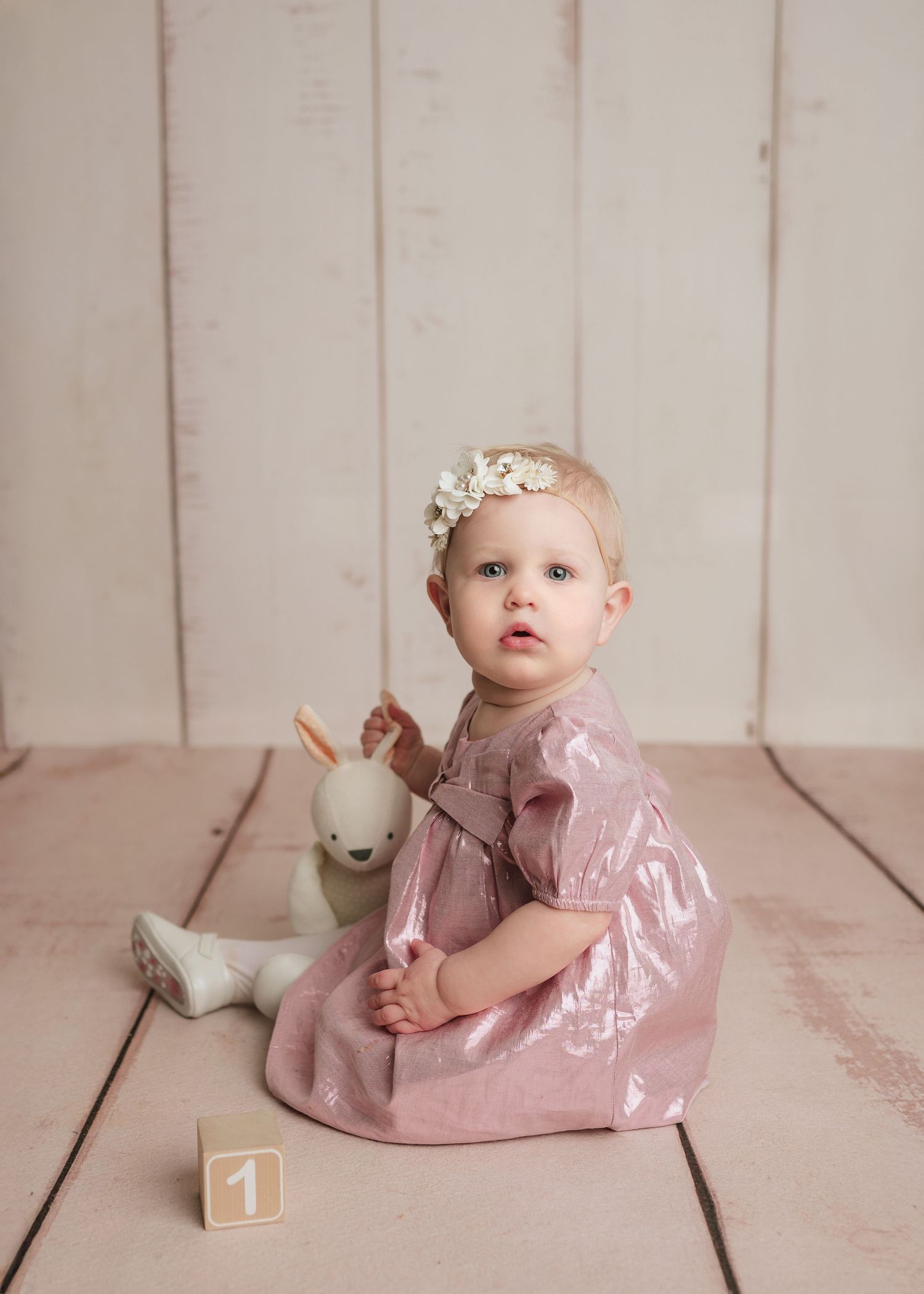 Baby girl in pink dress holding stuffed bunny toy with number one block on light wood floor.