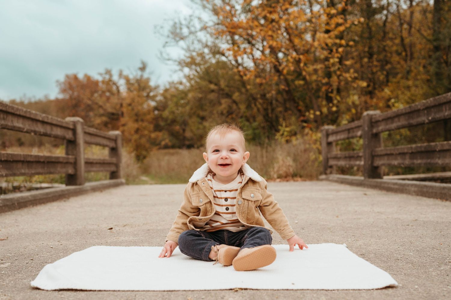 Smiling baby boy in tan jacket sitting on white blanket on wooden bridge with fall foliage.