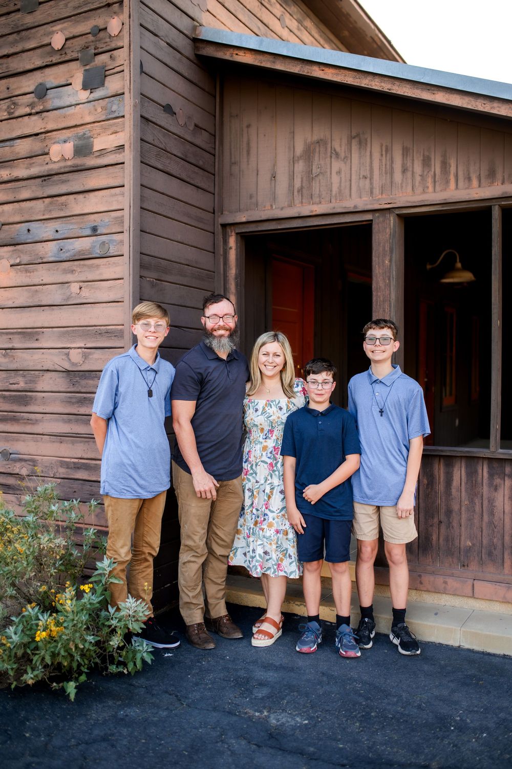 A smiling family of five poses outside a rustic wooden building, featuring two adults and three boys in casual attire.