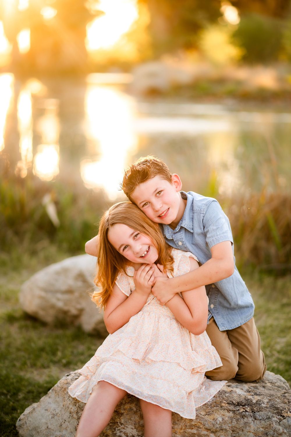 Two smiling children hugging on a rock at golden hour near a pond, boy in blue shirt embracing girl in pink dress.