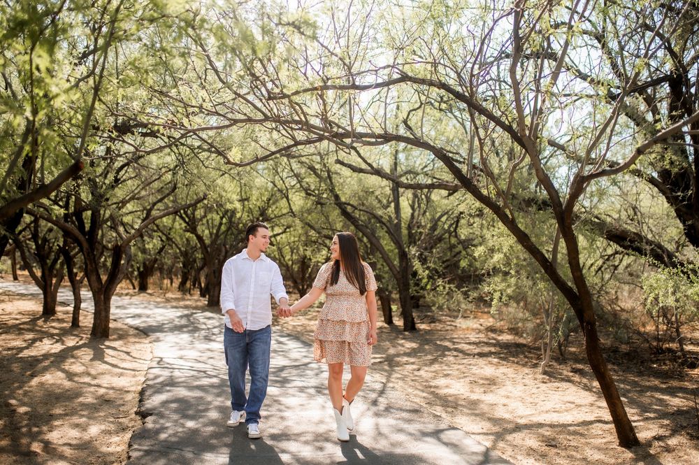 Couple holding hands and walking on a sunny tree-lined path during an outdoor engagement photo session.