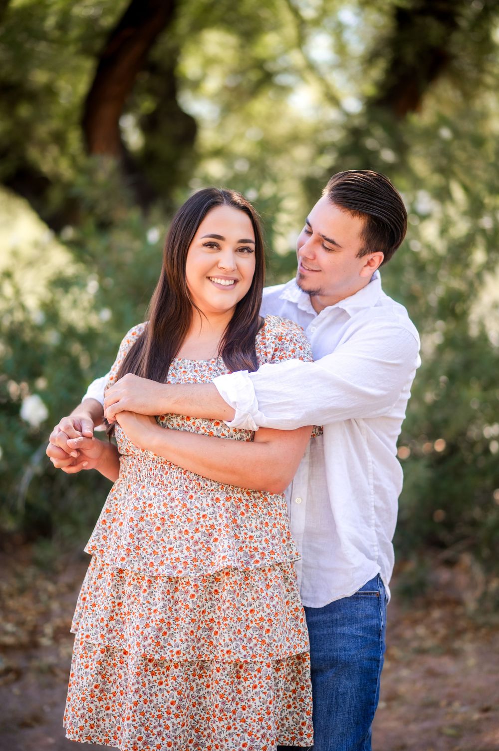 Couple embracing outdoors, woman in floral dress smiling at camera, man in white shirt hugging her from behind among green trees.