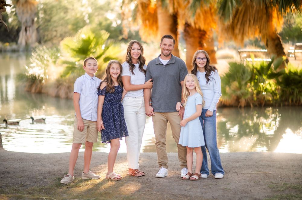 Family of six smiling outdoors near a pond with palm trees and ducks in the background on a sunny day.