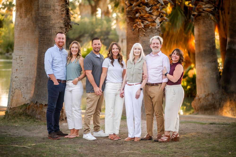 Seven smiling family members pose together outdoors among tall palm trees near a serene lake on a sunny day.