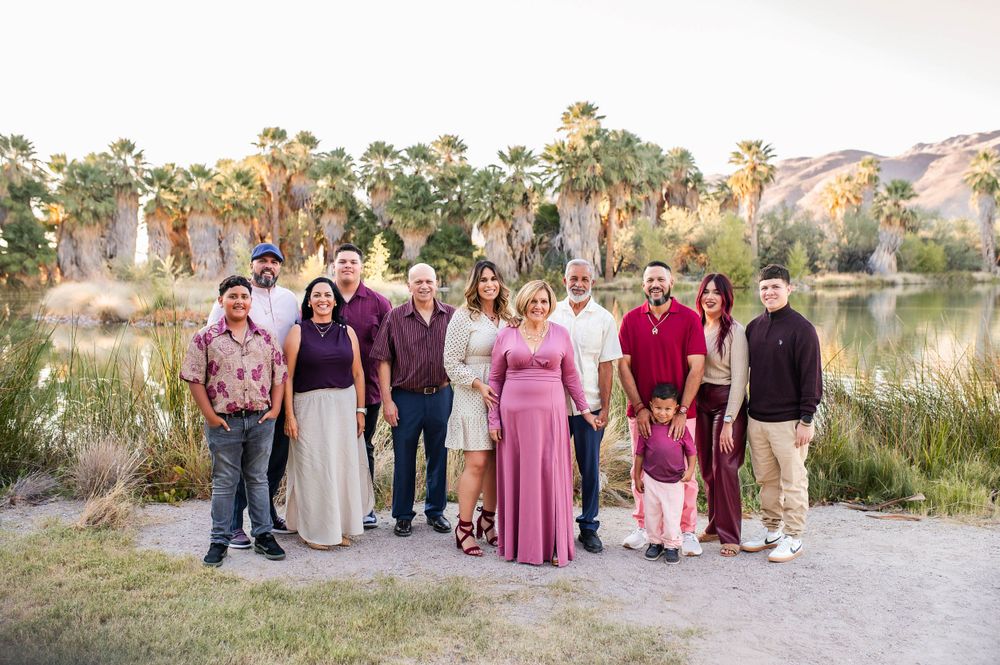 Large extended family portrait outdoors near a pond with lush palm trees at sunset.