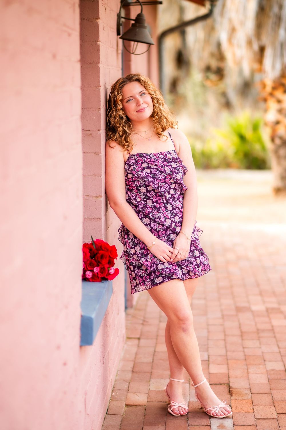 Young woman in purple floral mini dress and white heels leaning against pink wall holding red roses outdoors.