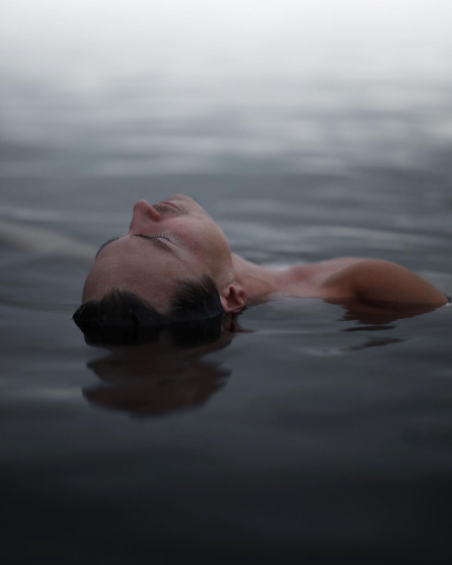 A person floats serenely in dark water with their head tilted back in a tranquil and moody composition.