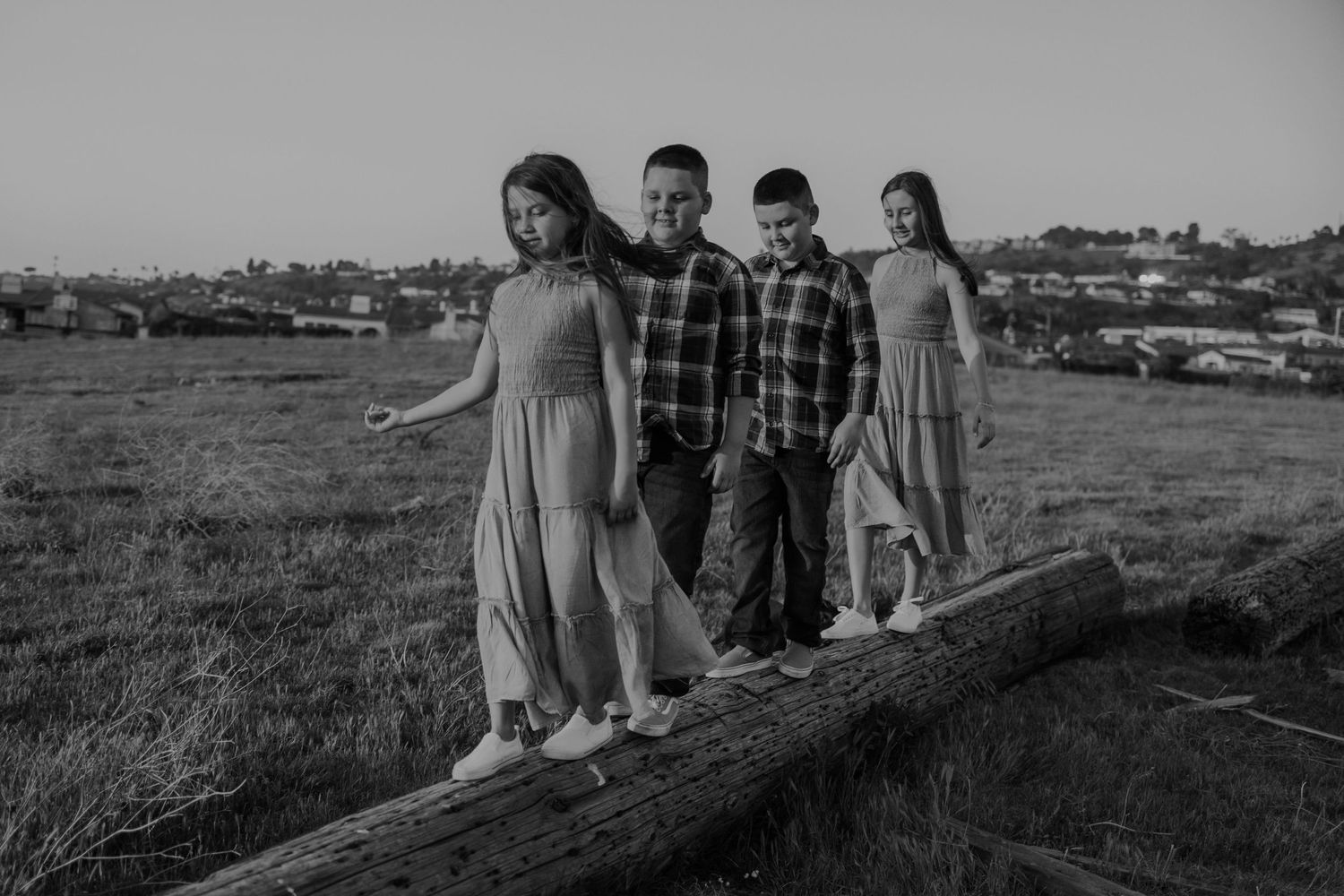 Four children walking on a large log in a grassy field with houses on a hillside in the background.