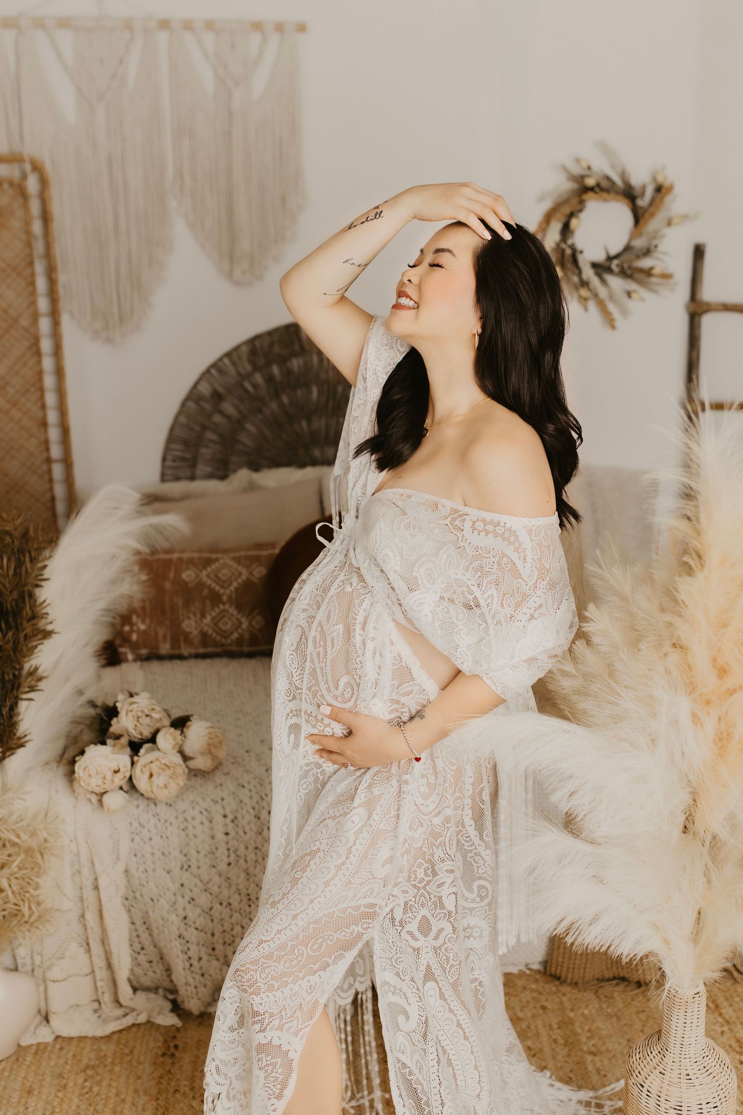 Expecting mother in white lace dress laughing joyfully in boho-styled room with dried flowers.