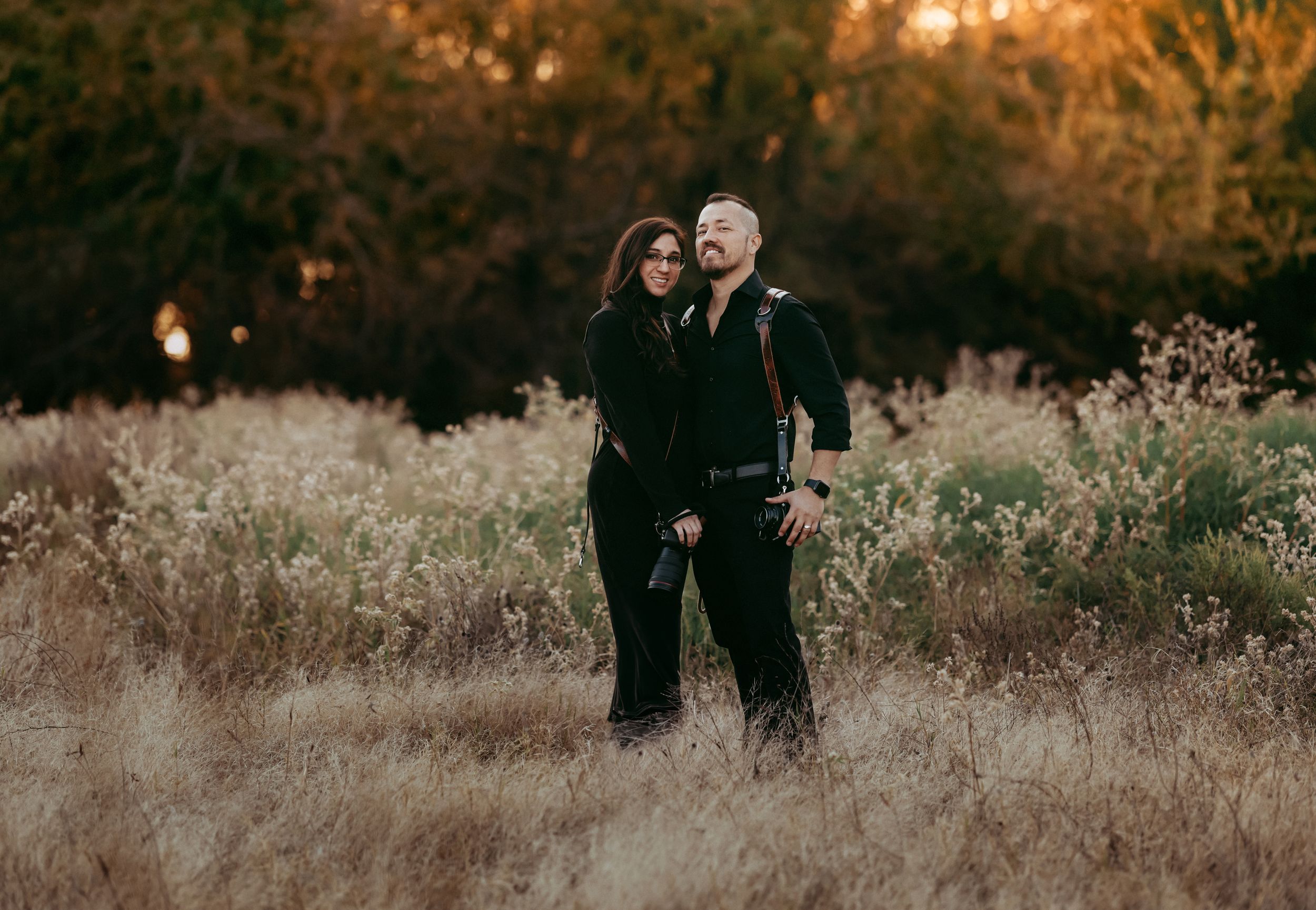 A couple dressed in black stands together in a golden field at sunset.