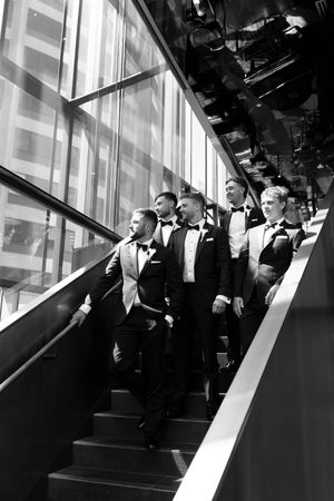 Groomsmen in tuxedos descend a modern staircase in a black and white wedding photo.