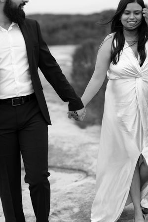 Black and white photo of a smiling bride in white gown holding hands with groom in black suit outdoors.