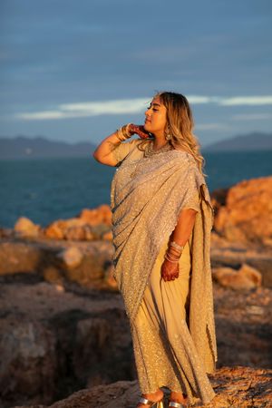 A person in a sparkly gold dress poses dramatically against a rocky coastal landscape at golden hour.