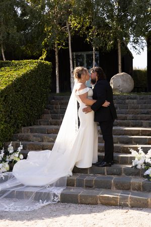 Elegant outdoor wedding ceremony on stone steps with flowing white veil and floral arrangements in garden setting.