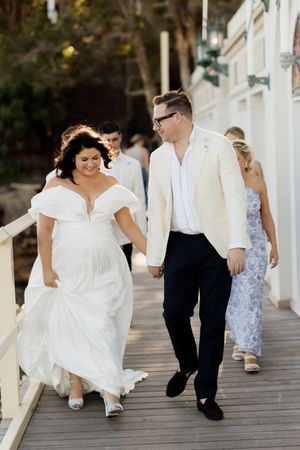 Happy couple walking on a wooden boardwalk during a beach wedding, with the bride wearing a flowing white off-shoulder gown.