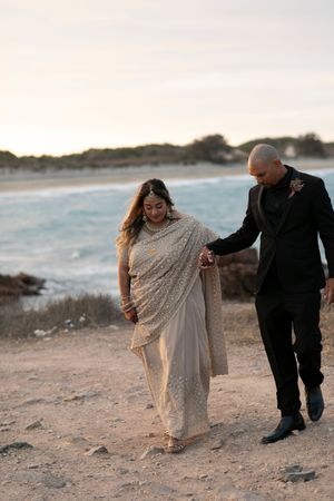 Couple in traditional Indian attire walks hand-in-hand along a sandy beach at sunset near turquoise ocean waves.