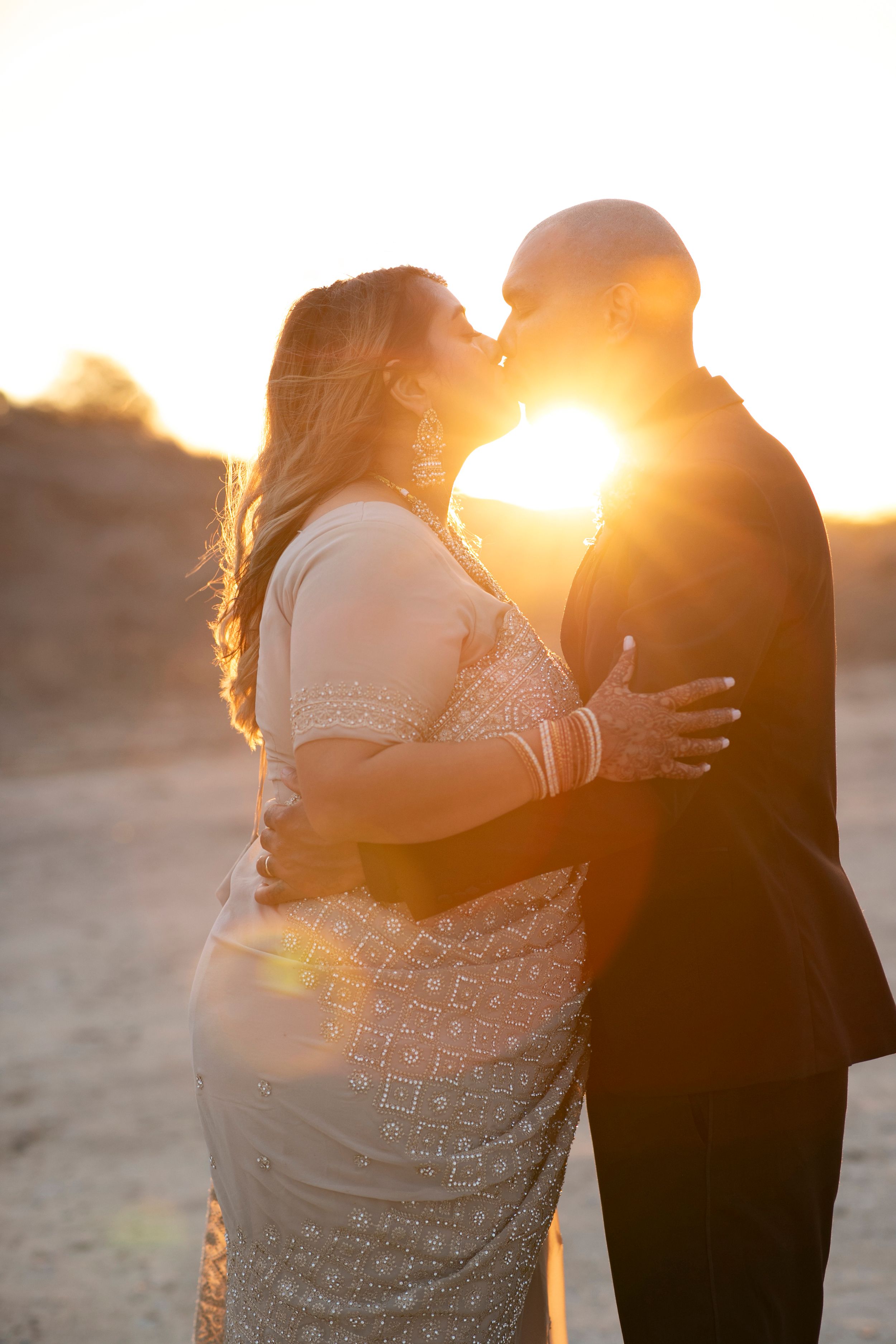 Couple sharing a romantic kiss at golden sunset, woman in embroidered sari, man in dark suit, warm backlit glow.