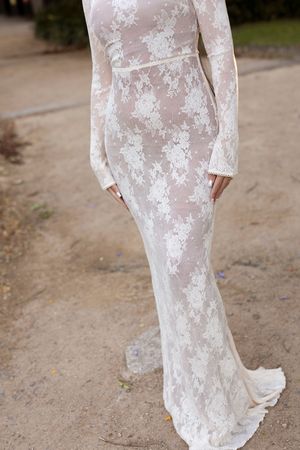 Elegant white lace long-sleeve wedding gown with floral pattern and fitted silhouette, photographed outdoors.