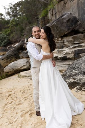 A happy bride and groom embrace on a sandy beach surrounded by rocky cliffs on their wedding day.